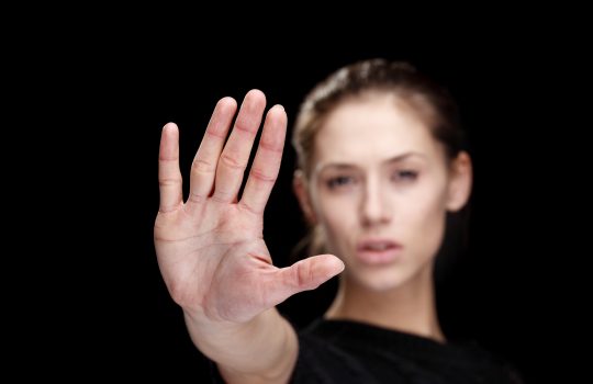Woman signaling STOP. Headshot of a woman looking straight at the camera, posing in studio, holding up her hand, black background. Focus on hand.